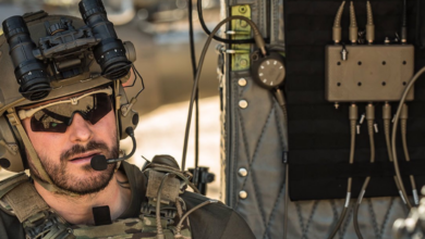 A soldier is seen beside INVISIO's Intercom system, a rectangular device with eight ports on its top and bottom sides. The soldier, equipped in camouflage, also has a headset, a pair of binoculars, and a radio.