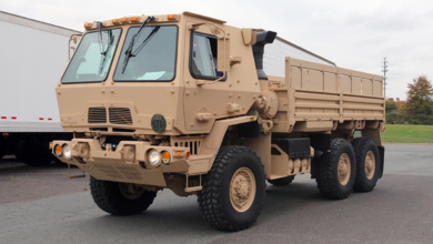 A brown M1083 army truck is seen parked in what appears to be a parking area. The six-wheel truck has two windshields and an open-trunk back.