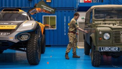 A British Army land rover with an electric engine is seen parked on the right side of the image, right beside a NEOM McLaren Extreme E racecar on the left. A uniformed army personnel is seen reaching for the land rover's door, as if opening it. The rover is painted brown-green, with a plate in its front that reads "LURCHER".The racecar is painted black with orange decals.