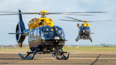 Two Airbus H145 helicopters are seen operating in what appears to be an aircraft runway. The helicopters are painted black, with its rotary blades components and base painted yellow. The one in the foreground has the number "98" painted in white in front of its cockpit, while the one in the back has "97".