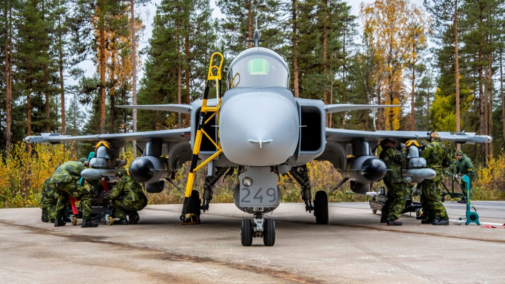 A Gripen C-series plane is seen being serviced by uniformed personnel while parked in the ground. All seven personnel are wearing green camouflage uniforms. The plane is painted gray with the number "249" painted in darker gray right below the front of the cockpit. The background is a forested area with trees and shrubbery.
