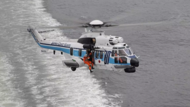A Japan Coast Guard H225 helicopter is seen flying above gray waters. The white aircraft has a thin navy blue and thick light blue stripe painted on its side, as well as "JAPAN COAST GUARD" written on its tail boom. Three personnel can be seen inside the helicopter: one is the pilot inside the cockpit, and the other two are on the passenger seats.