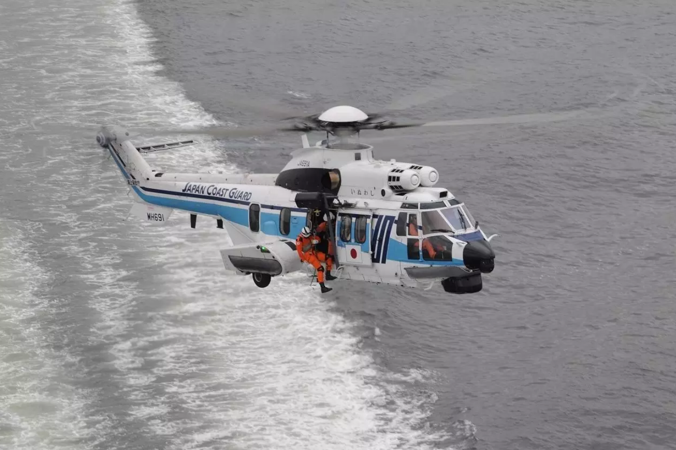 A Japan Coast Guard H225 helicopter is seen flying above gray waters. The white aircraft has a thin navy blue and thick light blue stripe painted on its side, as well as "JAPAN COAST GUARD" written on its tail boom. Three personnel can be seen inside the helicopter: one is the pilot inside the cockpit, and the other two are on the passenger seats.