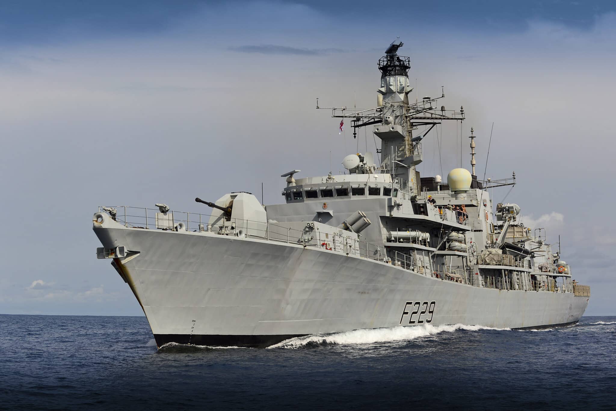 UK's HMS Lancaster warship is seen sailing deep blue waters. The gray ship has "F229" painted on its side in black. The background is a dull blue sky covered by dark clouds.