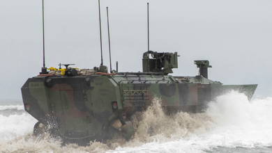 An Amphibious Combat Vehicle is seen trudging on ocean waters. The vehicle has four antennae on top of it, all pointing straight up. It's painted in camouflage. A misty mix of white and muddy brown water surrounds it as it traverses the waters.