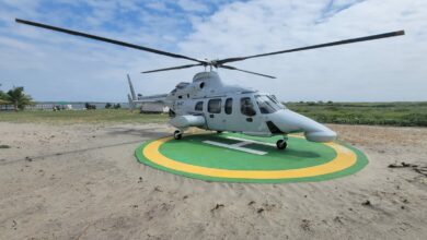 The Ecuadorian Navy's Bell 430 aircraft with the ID HN-407 is seen on a helipad in the middle of a sandy area. The helicopter is painted white, with "HN-407" written on the right side of its body in black. On the back, green grass can be seen stretching all the way to the horizon. A body of water peeks a little bit farther. The sky is blue with lots of thin white clouds.
