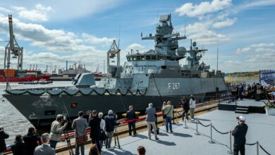 The Karlsruhe, a German Navy corvette, is seen docked in a pier during its christening ceremony. The gray ship's deck railings are decorated with a ribbon made of leaves. The middle of its hull has "F267" painted on it in white. A group of journalists, photographers, and navy officers and personnel are positioned on the dock, observing the corvette. The background is a clear blue sky with some light white clouds.