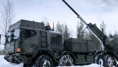 A Rheinmetall HX truck is seen parked on a snowy field. The grayish-green 8x8 truck has chains fitted on some of its tires to help with traction. On its back, a large turret is attached and pointing northwest. The ground is covered completely by snow, covering almost the entire bottom half of the truck's wheels. The background is a sparse group of thin trees with little to no leaves, and a gloomy, white sky.