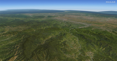 A shot of a land mass generated by Precision3D Data Suite by Maxar Intelligence. The computer-generated image shows a relatively flat and green landscape with small mountains. The background is a clear blue sky.