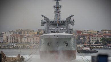 The Logistics Support Ship Atlante is seen floating in a shipyard's waters. Most of the ship is painted gray; its bottom part is painted red. Blurred and cut out figures of onlookers are seen on the foreground. The background is a gray, foggy sky serving as the backdrop for a horizon filled with what appears to be residential complexes and a number of boats in the water.