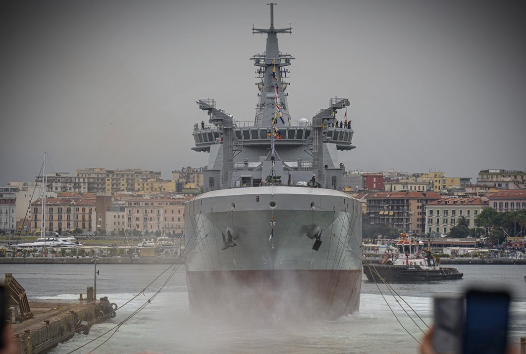 The Logistics Support Ship Atlante is seen floating in a shipyard's waters. Most of the ship is painted gray; its bottom part is painted red. Blurred and cut out figures of onlookers are seen on the foreground. The background is a gray, foggy sky serving as the backdrop for a horizon filled with what appears to be residential complexes and a number of boats in the water.