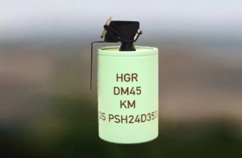 A Rheinmetall DM 45 hand grenade is displayed. The grenade has a cylindrical shape and is painted green, with "HGR DM45 KM" and a long serial number inscribed on it.