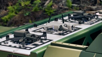 Three aerial drones are seen stationed and charging on a Drone Swarm Tactical OverWatch platform. The drones are charging via wireless technology. The middle drone is painted white, while the two flanking it are painted black.