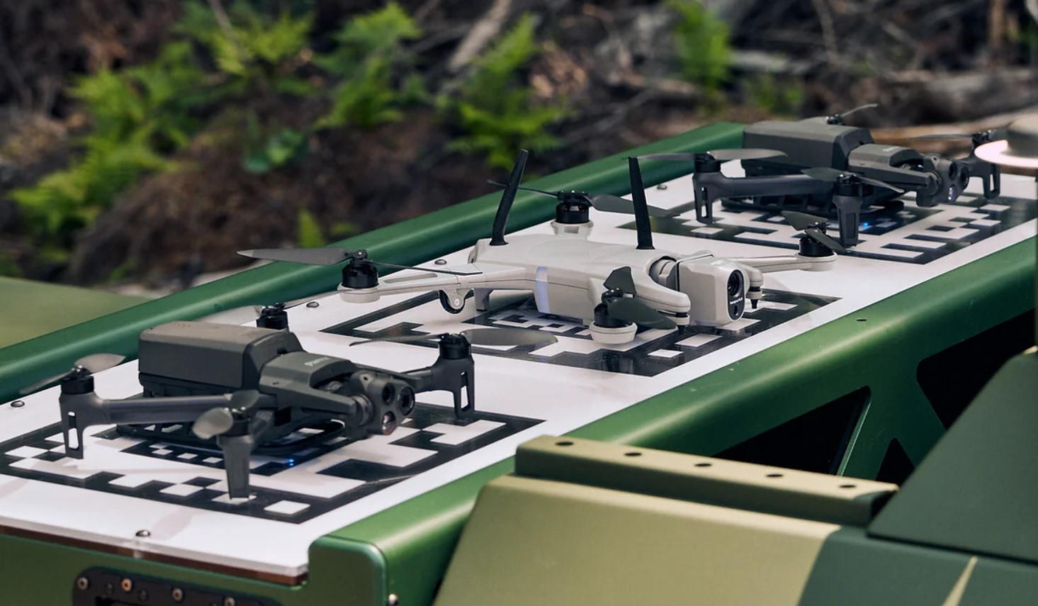 Three aerial drones are seen stationed and charging on a Drone Swarm Tactical OverWatch platform. The drones are charging via wireless technology. The middle drone is painted white, while the two flanking it are painted black.