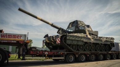 A Leopard 2A7HU battle tank is seen loaded on the back of a truck. The tank is painted in camouflage.