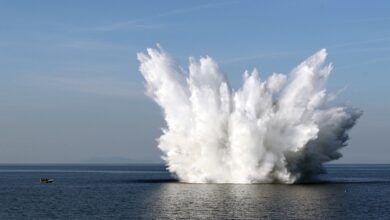 A naval mine sets off in the middle of an ocean. The water's white mist is seen blasting off on all directions. On the far left side of the image, a small boat is seen.