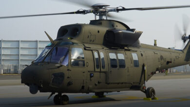 A KUH-1 Surion Helicopter is seen grounded in what appears to be a large open area, most likely a testing facility or a military headquarter. The aircraft is painted dark green, and its propellers and rotor blades are blurry as they spin.
