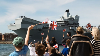 UK Royal Navy's HMS Prince of Wales is seen leaving Portsmouth Naval Base. 11 people bid farewell in the foreground, waving UK flags.