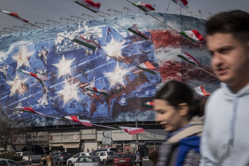 Iranian youths walk past a state building covered with a giant anti-US billboard depicting a symbolic image of the destroyed USS Abraham Lincoln (CVN-72) aircraft carrier in downtown Tehran, Iran, on January 28, 2026.
