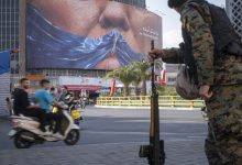 An armed Iranian police officer holding a rifle monitors the area as motorcyclists ride beneath a billboard depicting an AI-generated image of the Strait of Hormuz and an effigy of US President Donald Trump, displayed on the wall of a state building in downtown Tehran, Iran, on May 3, 2026.